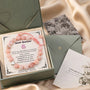 A pink beaded birthday bracelet with a small silver heart charm, presented in a sage green gift box with a handwritten message card. The box is styled on a beige satin fabric alongside a floral card and a green envelope.